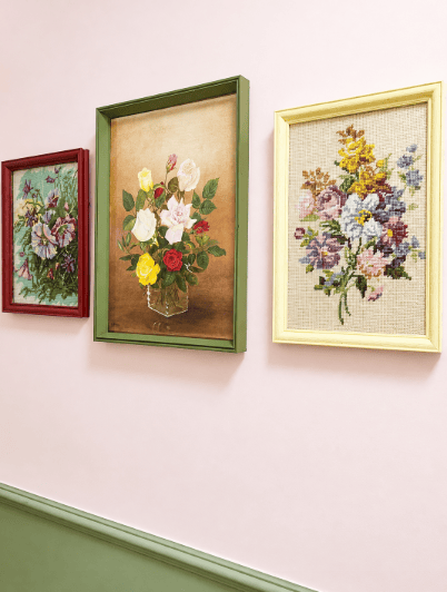 Three vintage floral needleworks in repainted green, maroon and yellow frames on a pink wall.